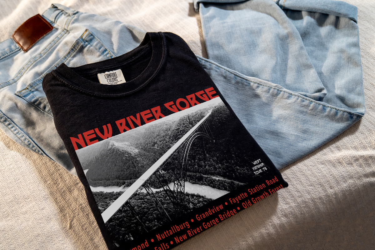 A black t-shirt with a red and white 'New River Gorge' design, laying on jeans