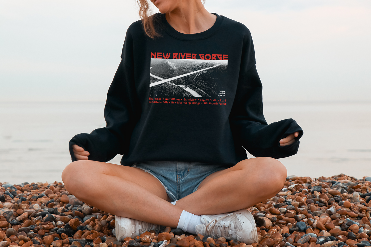 A person sitting on a beach wearing a black sweatshirt with 'NEW RIVER GORGE' printed on the front, paired with blue jean shorts and white sneakers.