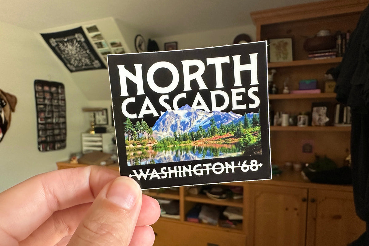 Person holding a 'North Cascades Washington '68' sticker in a room with furniture and decor.