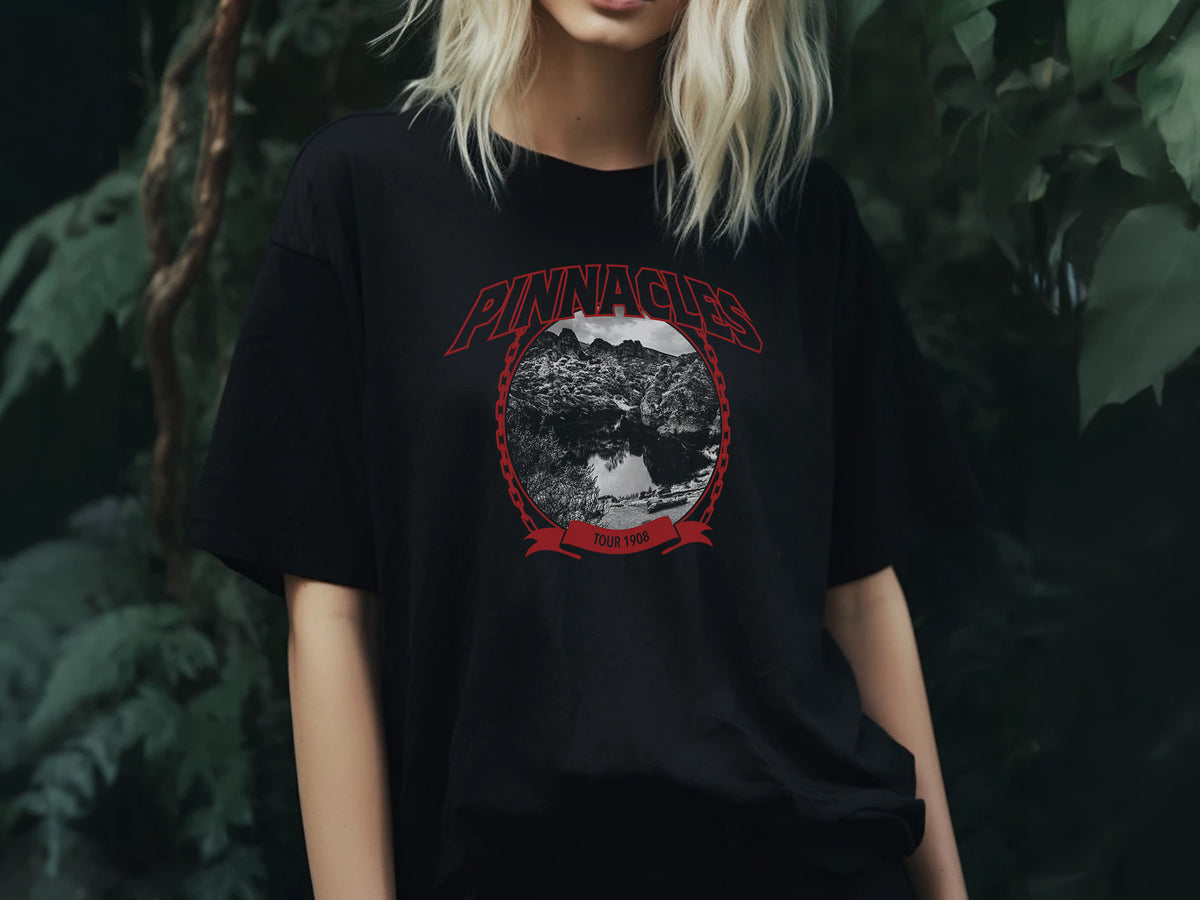 A person wearing a black t-shirt with a 'Pinnacles' design, standing in front of plants.