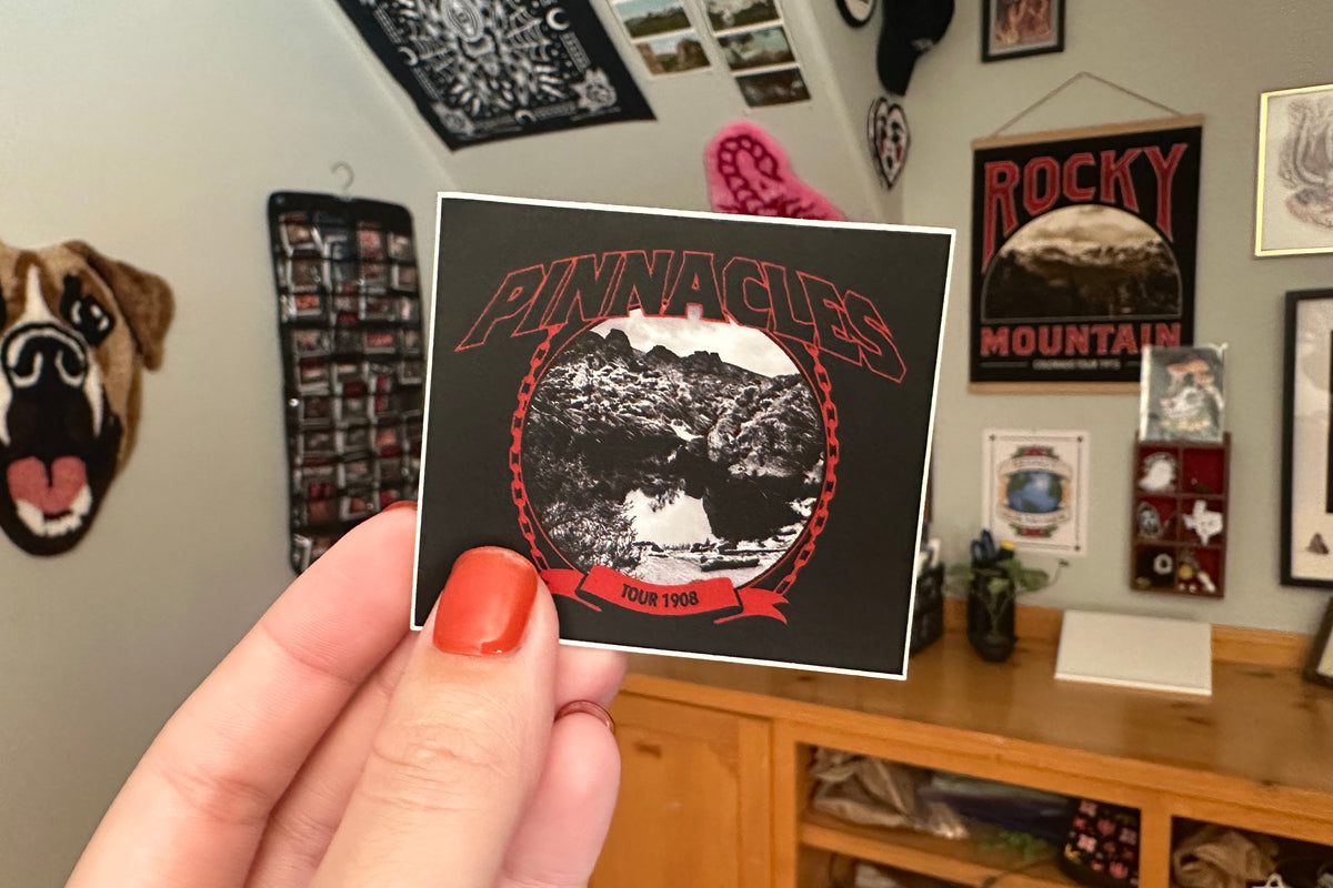 Hand holding a 'Pinnacles' sticker with a mountain design in a room with posters and decor.