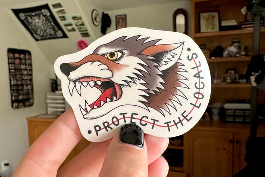 Sticker of a wolf with 'Protect the Locals' text held by a hand in a room.