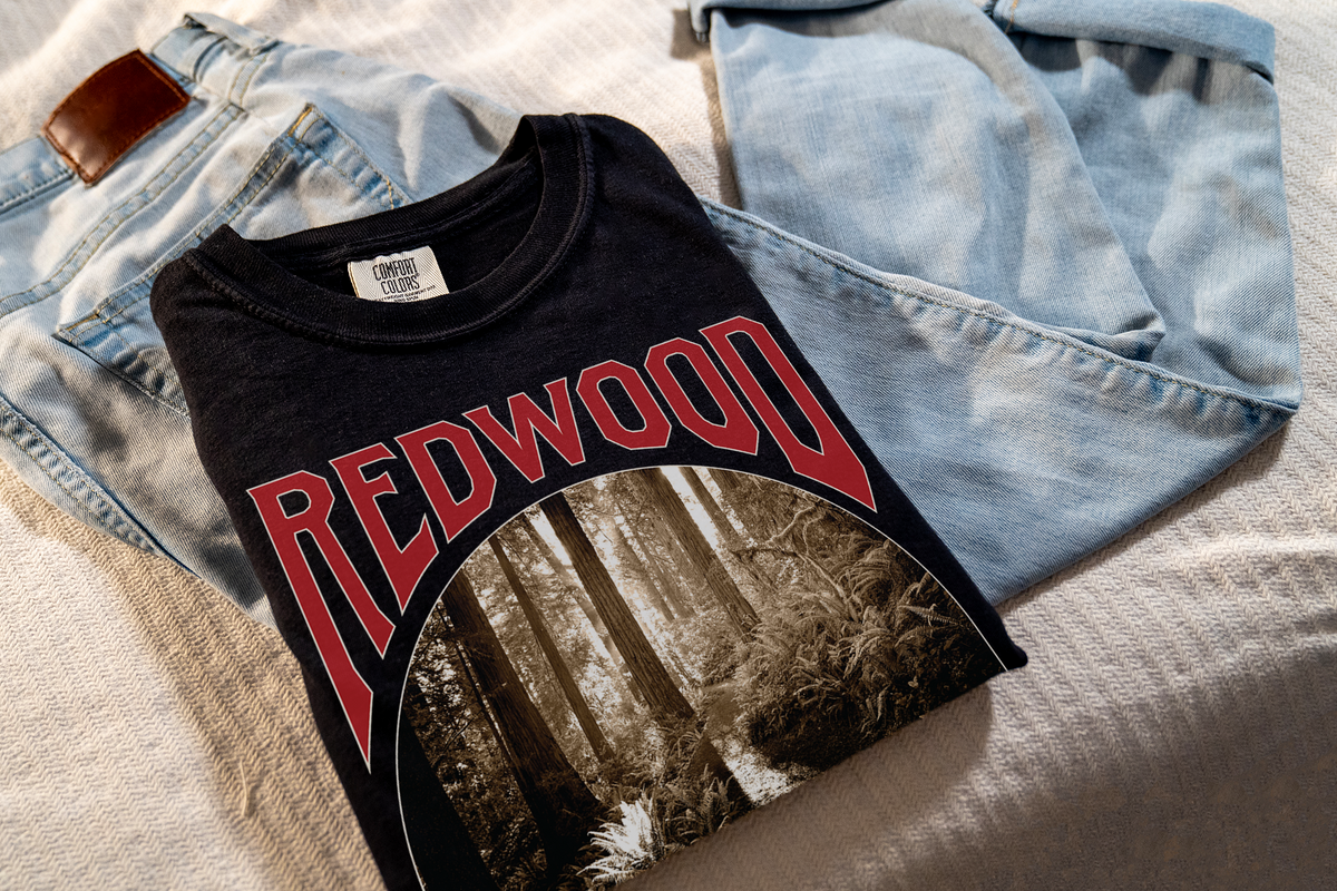 A black t-shirt with 'REDWOOD' written across the front, featuring a graphic of a redwood forest, laying on jeans.