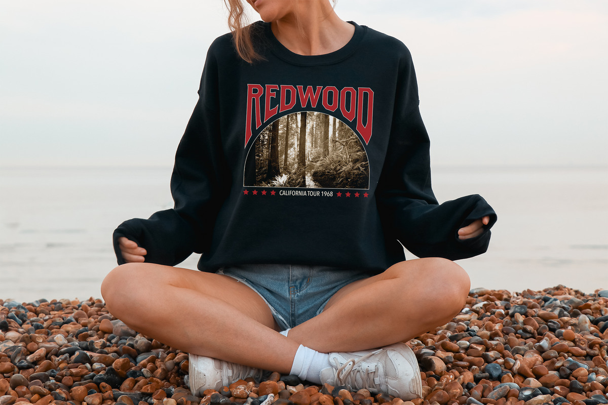 A person sitting on a pebble beach, wearing a black sweatshirt with a 'Redwood' graphic design.