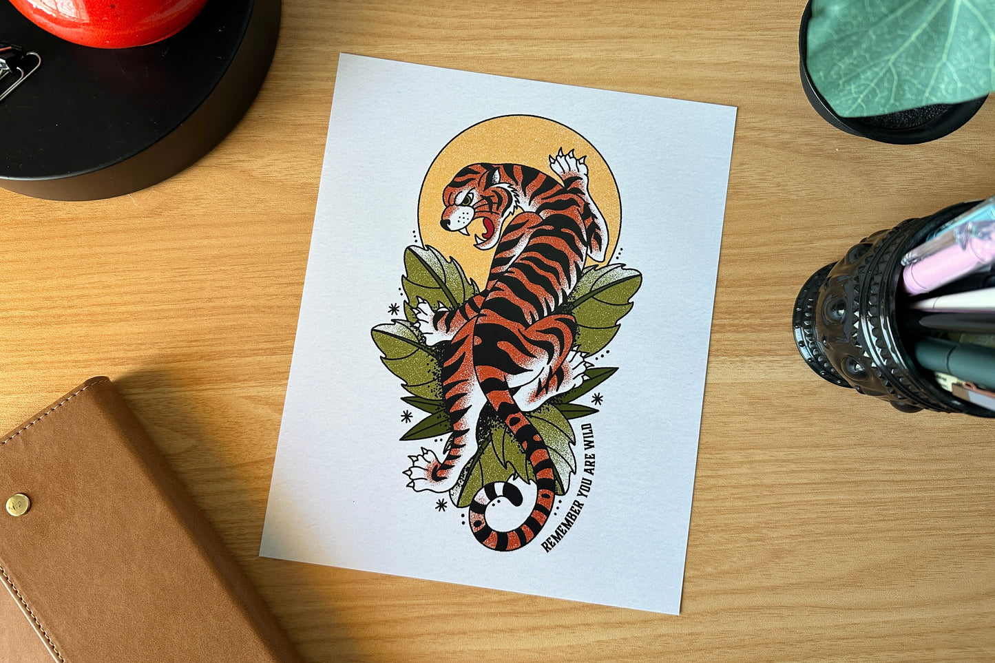 Tattoo design of a tiger on a white card with a pen holder and plant in the background.