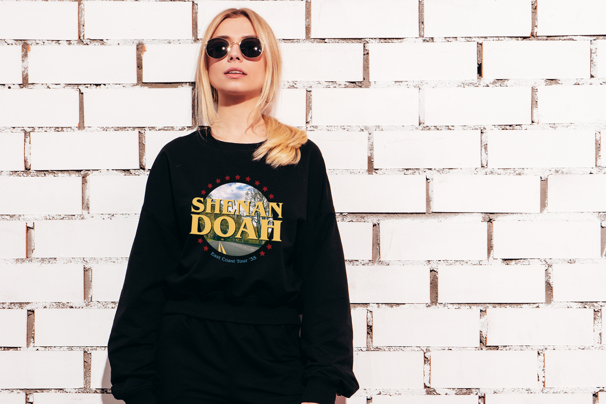 Person wearing a black sweatshirt with 'Shehan Doah' design against a white brick wall.