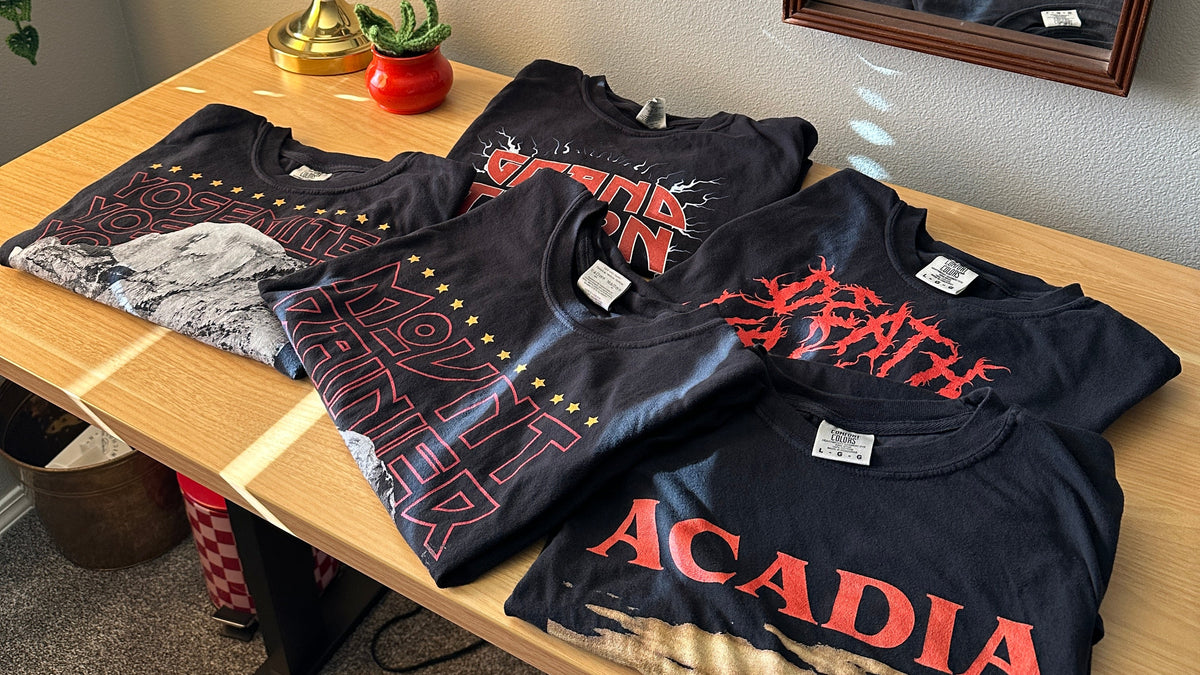 Several black graphic band t-shirts are laid out on a wooden desk in a room. 