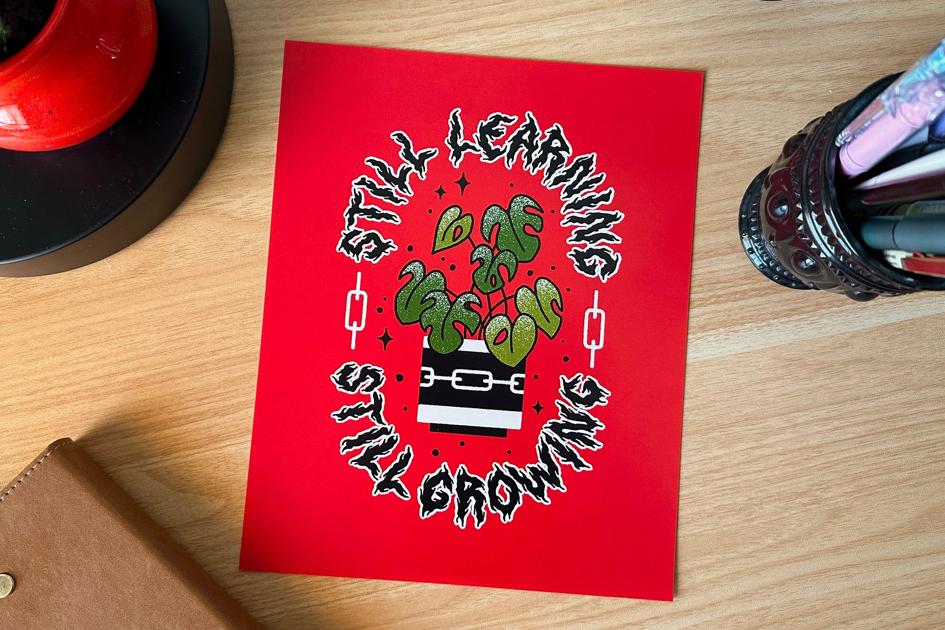 Red art print with green plant illustration and text on a wooden surface