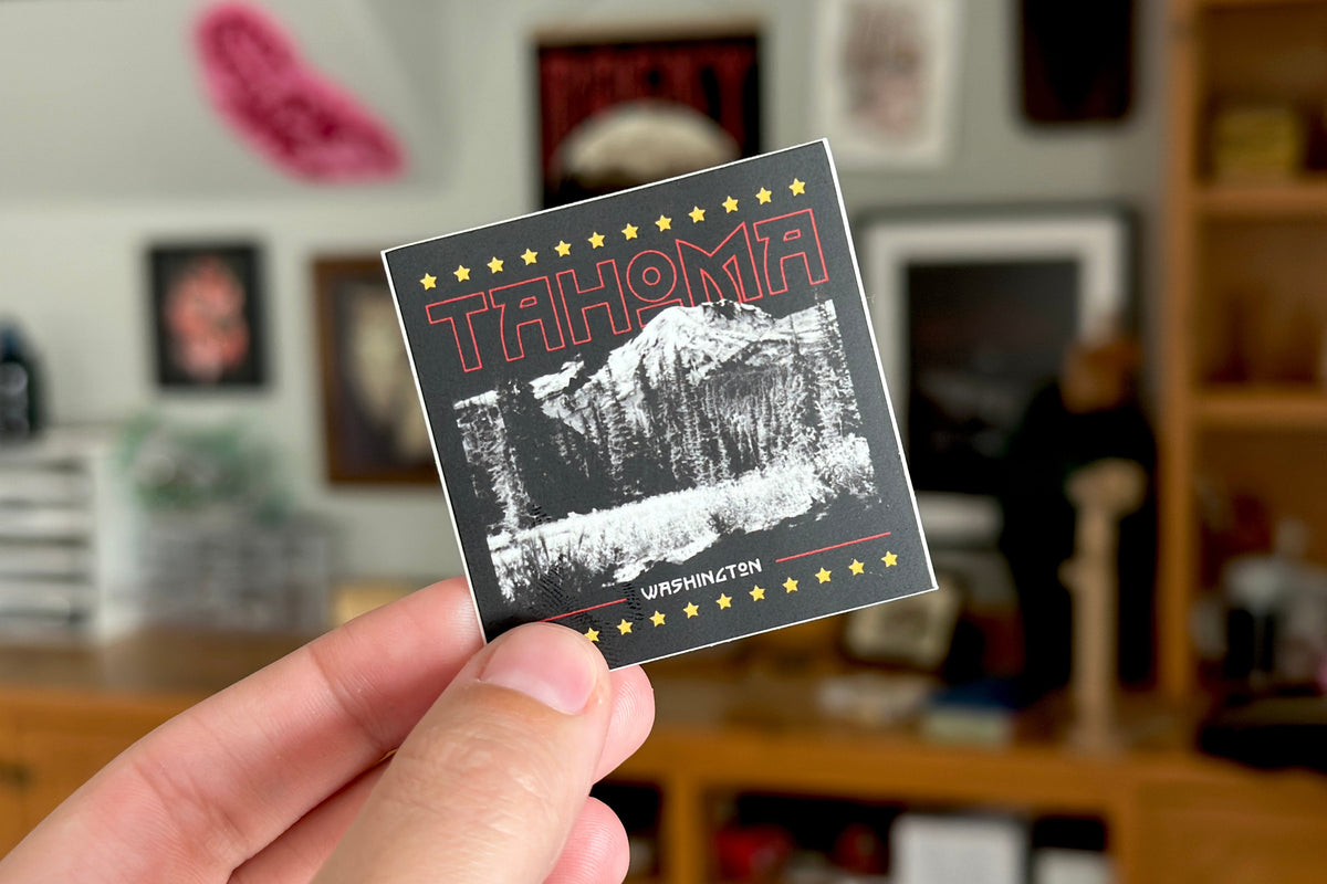 Hand holding a 'Tahoma Washington' sticker with a mountain graphic in a blurred indoor setting.