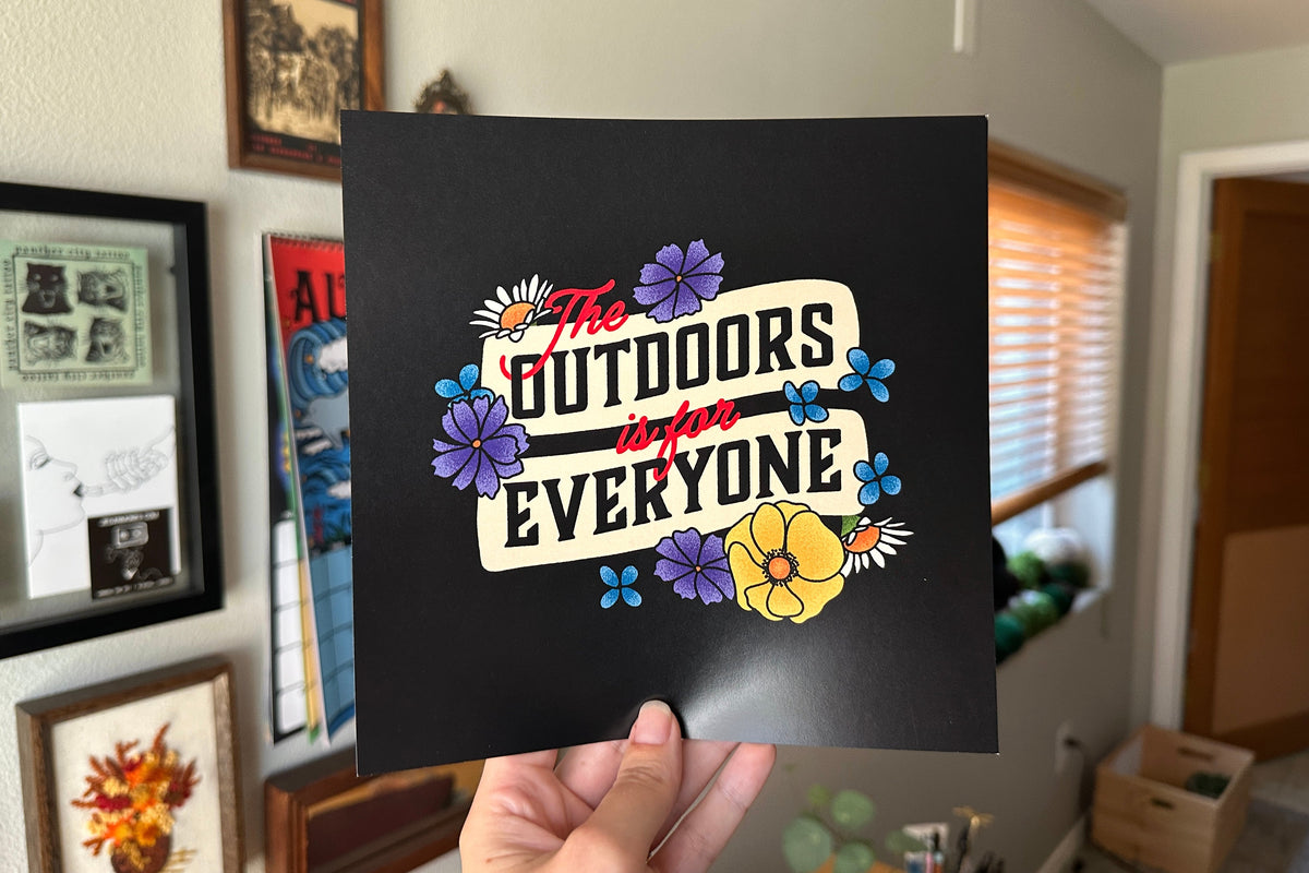 Hand holding a black sign with colorful text and flowers in a room with framed pictures on the wall.