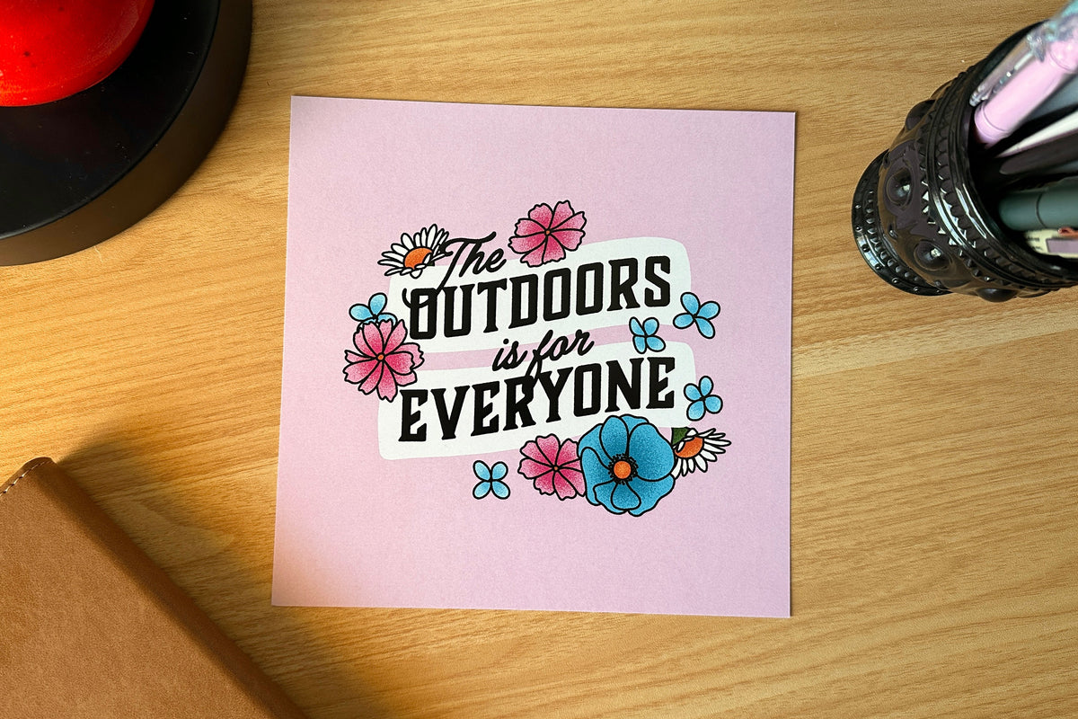 Art print with 'The Outdoors is for Everyone' text on a pink background, placed on a wooden surface.