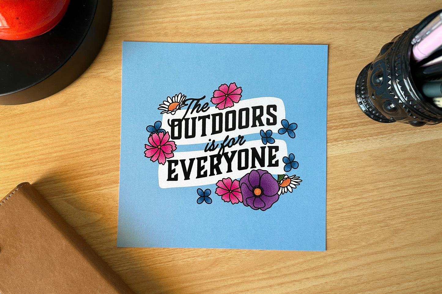 Art print with 'The Outdoors is for Everyone' text on a wooden surface with stationery items.