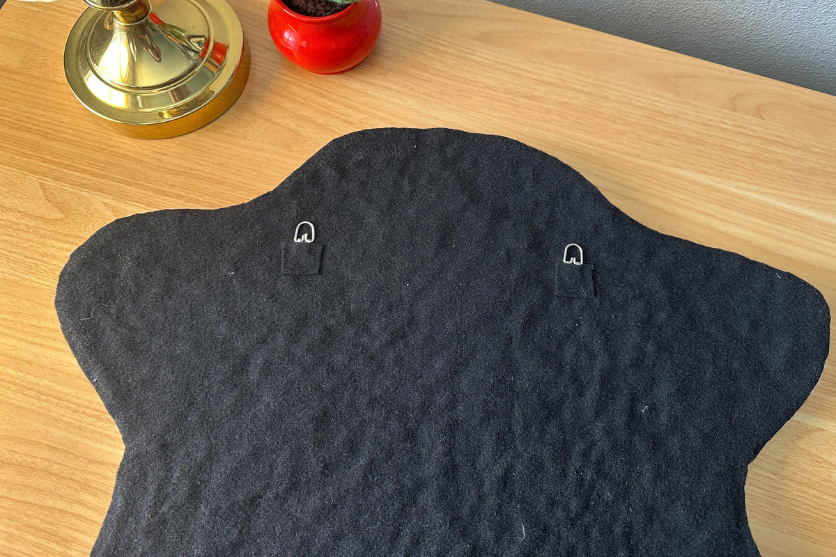 Black textured wall hanging on a wooden surface with a lamp and plant in the background.