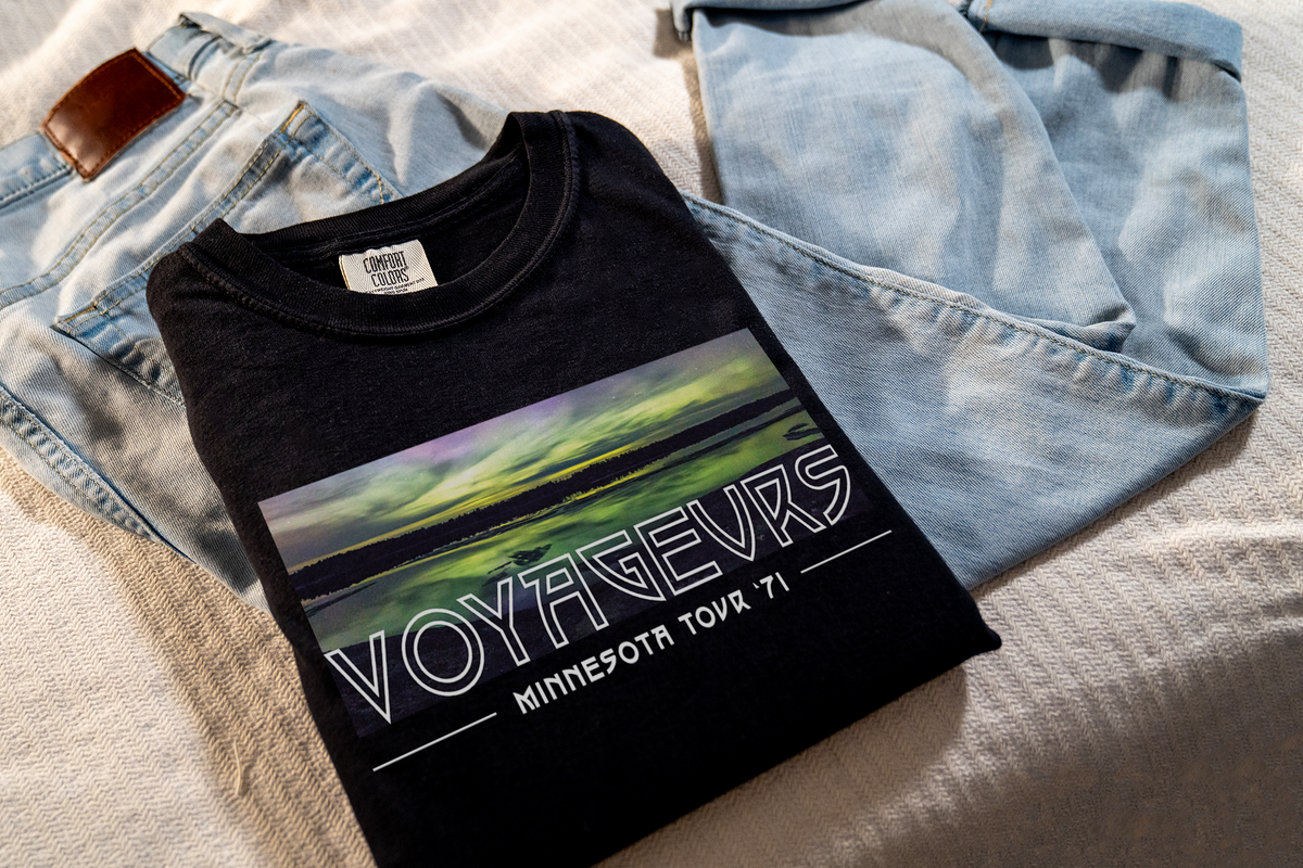 A black t-shirt with 'VOYAGEURS MINNESOTA TOUR 71' printed in white, displayed flat with a pair of blue jeans.