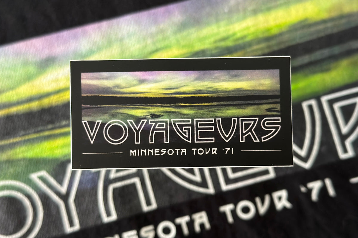 A black sticker with a 'Voyageurs' design, on a scenic background