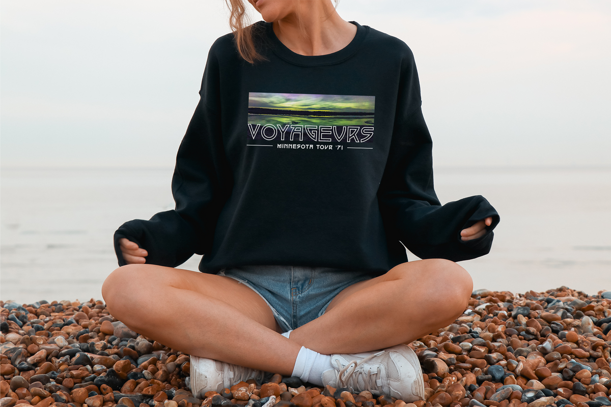 A person sitting on a pebble beach wearing a black sweatshirt with a 'Voyageurs' graphic design.