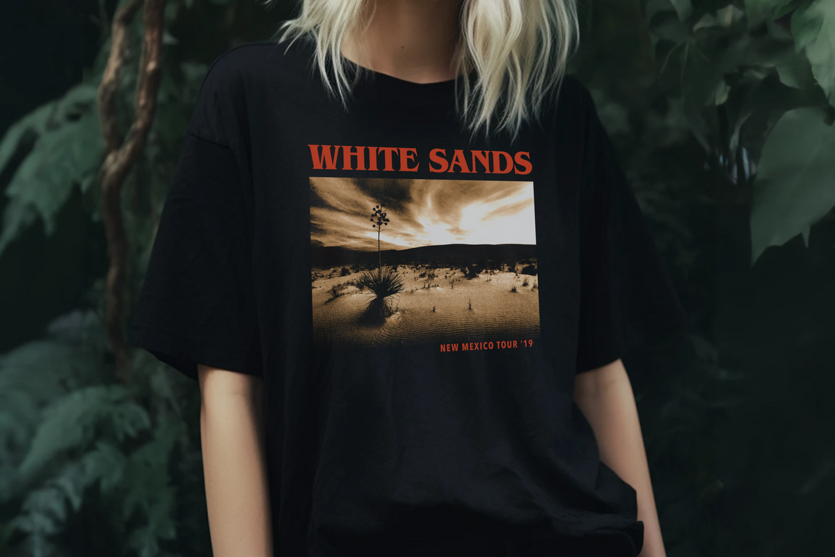 Person wearing a black t-shirt with 'White Sands' graphic design, standing in front of plants