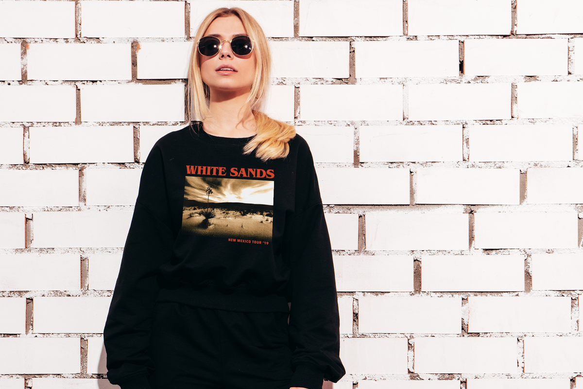 Person wearing a black sweatshirt with 'White Sands' design against a white brick wall.