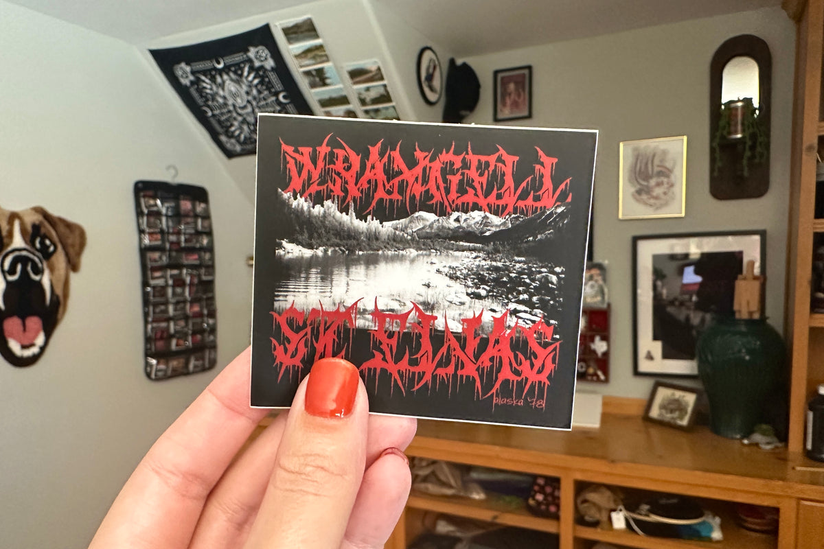 Person holding a 'Wrangell St Elias' sticker in a room with decor.