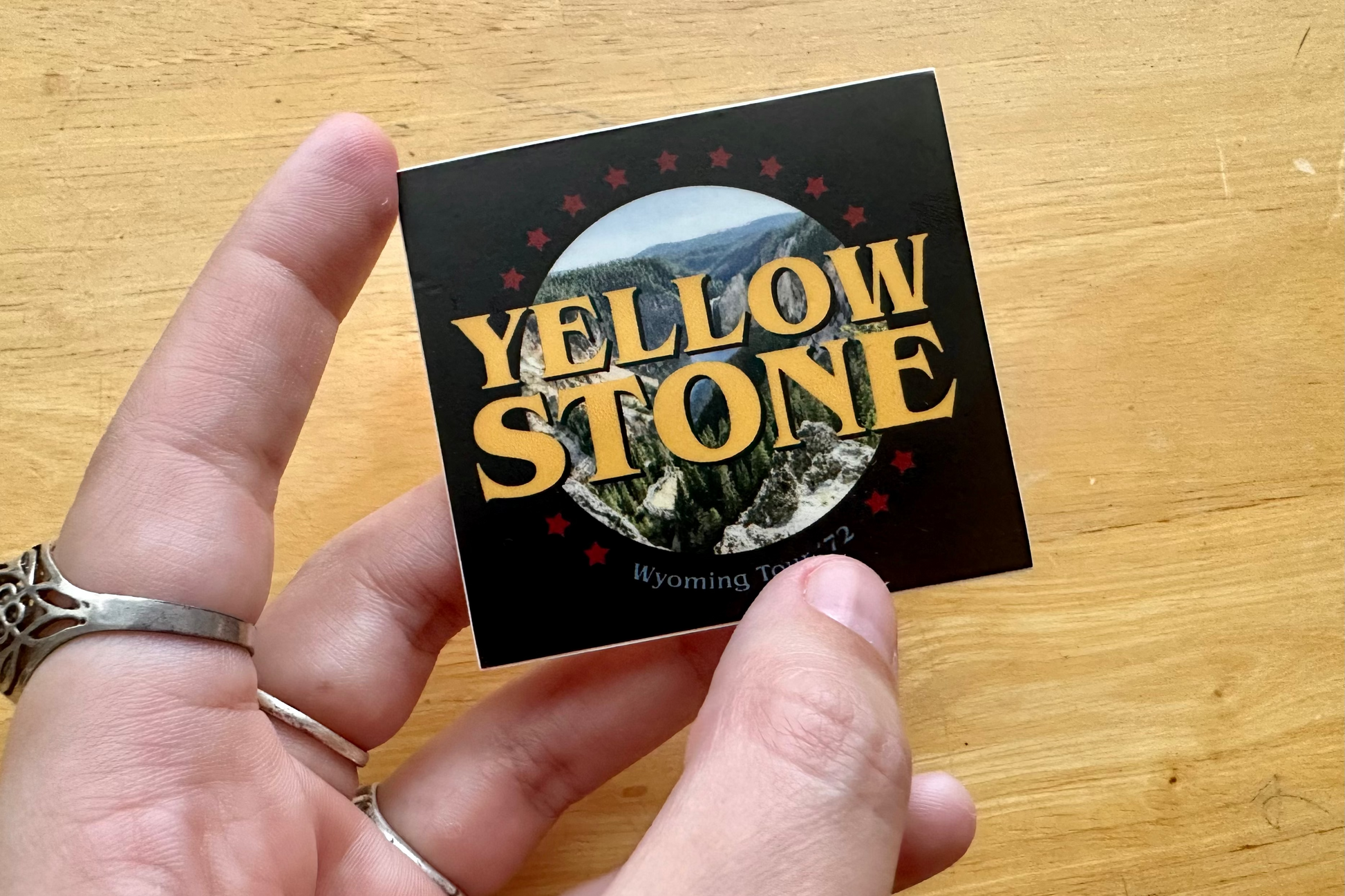 Hand holding a 'Yellowstone' sticker with a scenic image on a wooden surface