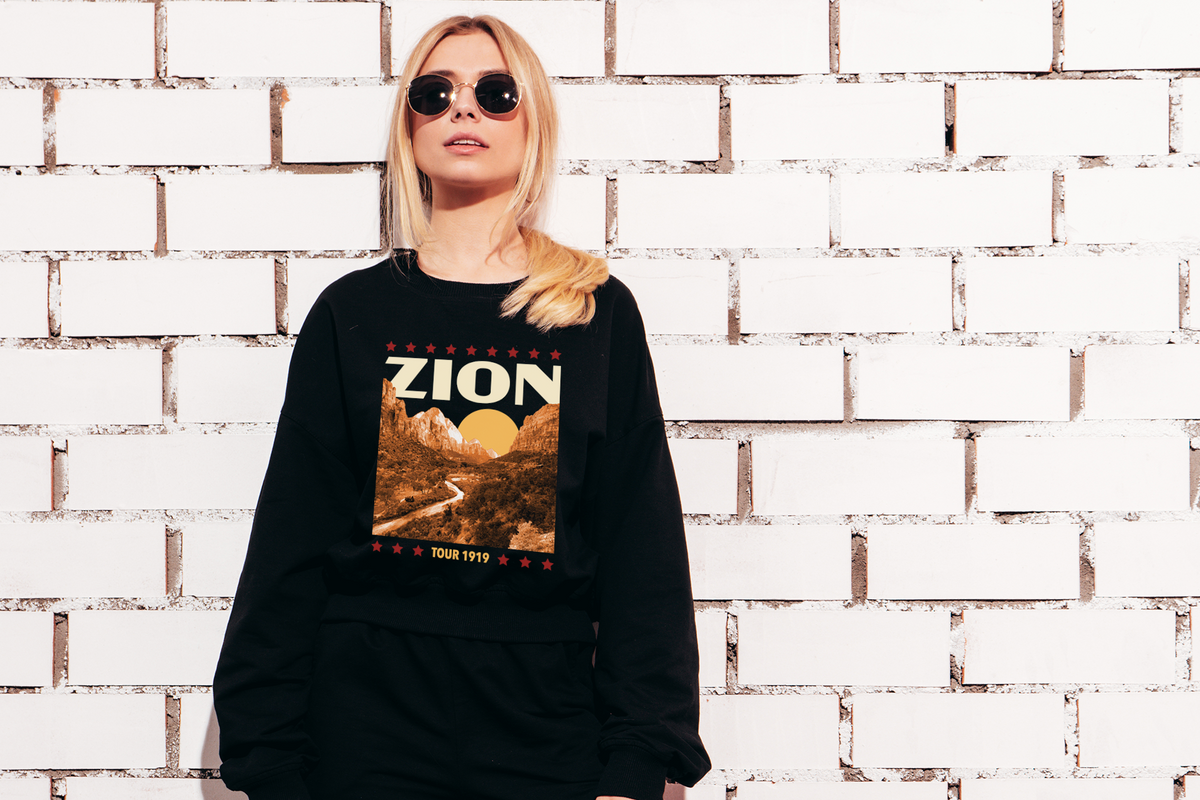 Person wearing a black sweatshirt with 'Zion' and mountain graphic against a white brick wall.